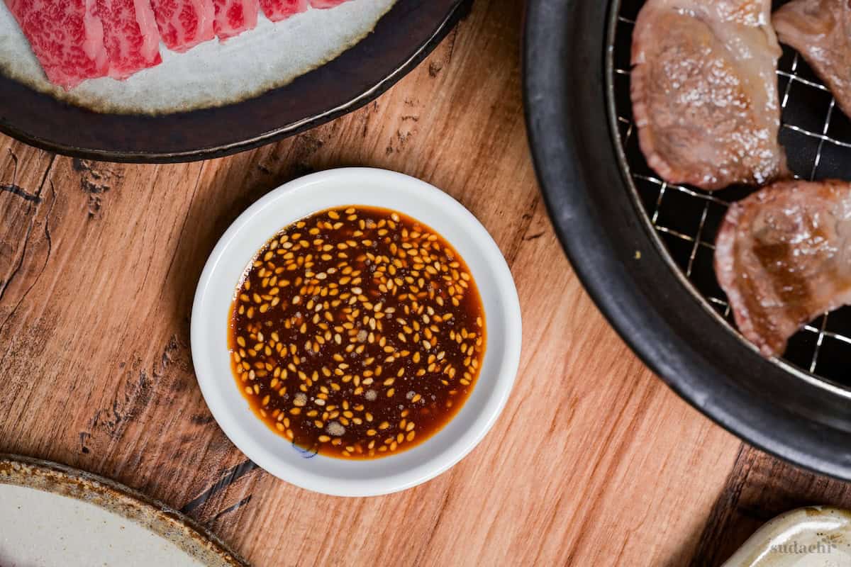 Homemade yakiniku sauce in a white bowl next to a plate of sliced raw beef, and a wire rack with grilled beef