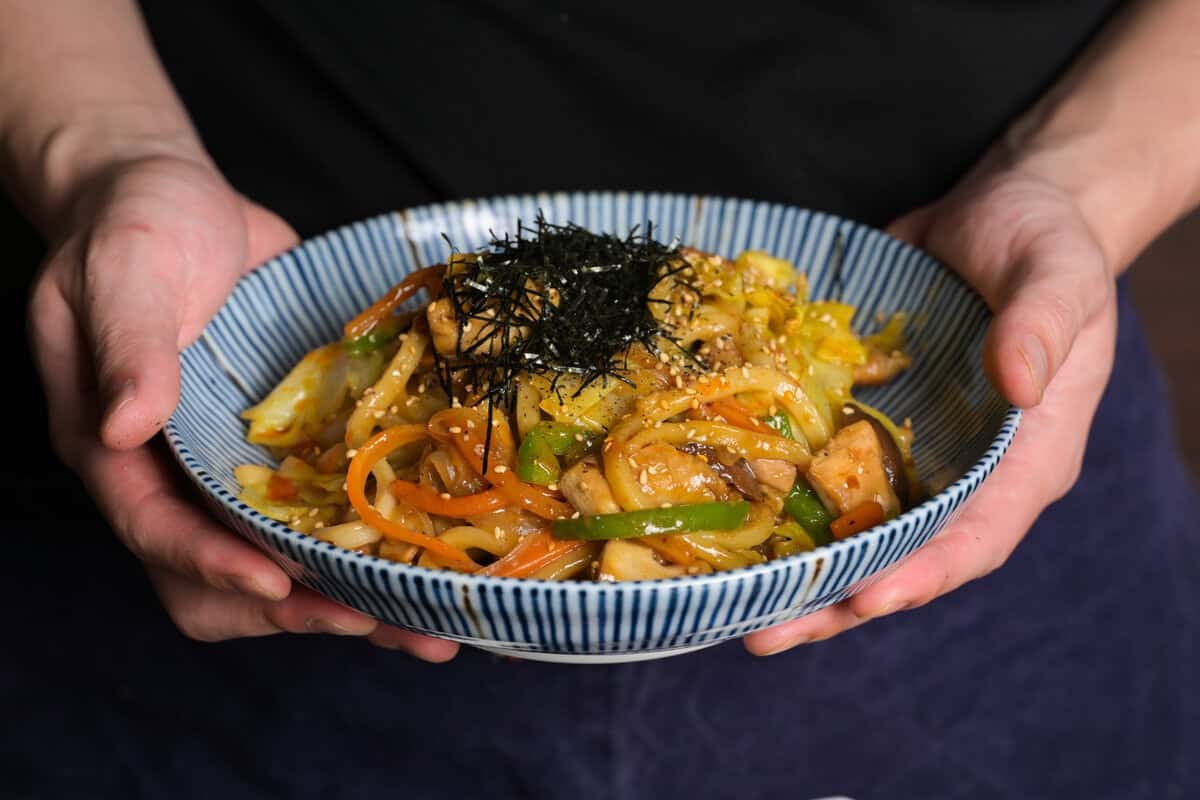 Hands holding a bowl of chicken yaki udon topped with shredded nori and sesame seeds, featuring stir-fried udon noodles with chicken, cabbage, carrots, and green peppers.