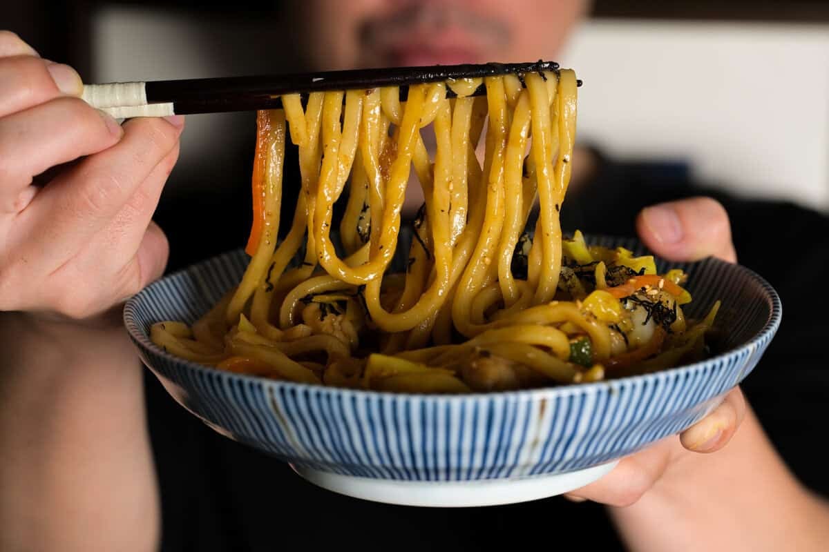 Chicken yaki udon noodles lifted with chopsticks from a blue striped bowl, showing glossy stir-fried udon with vegetables and savory sauce.