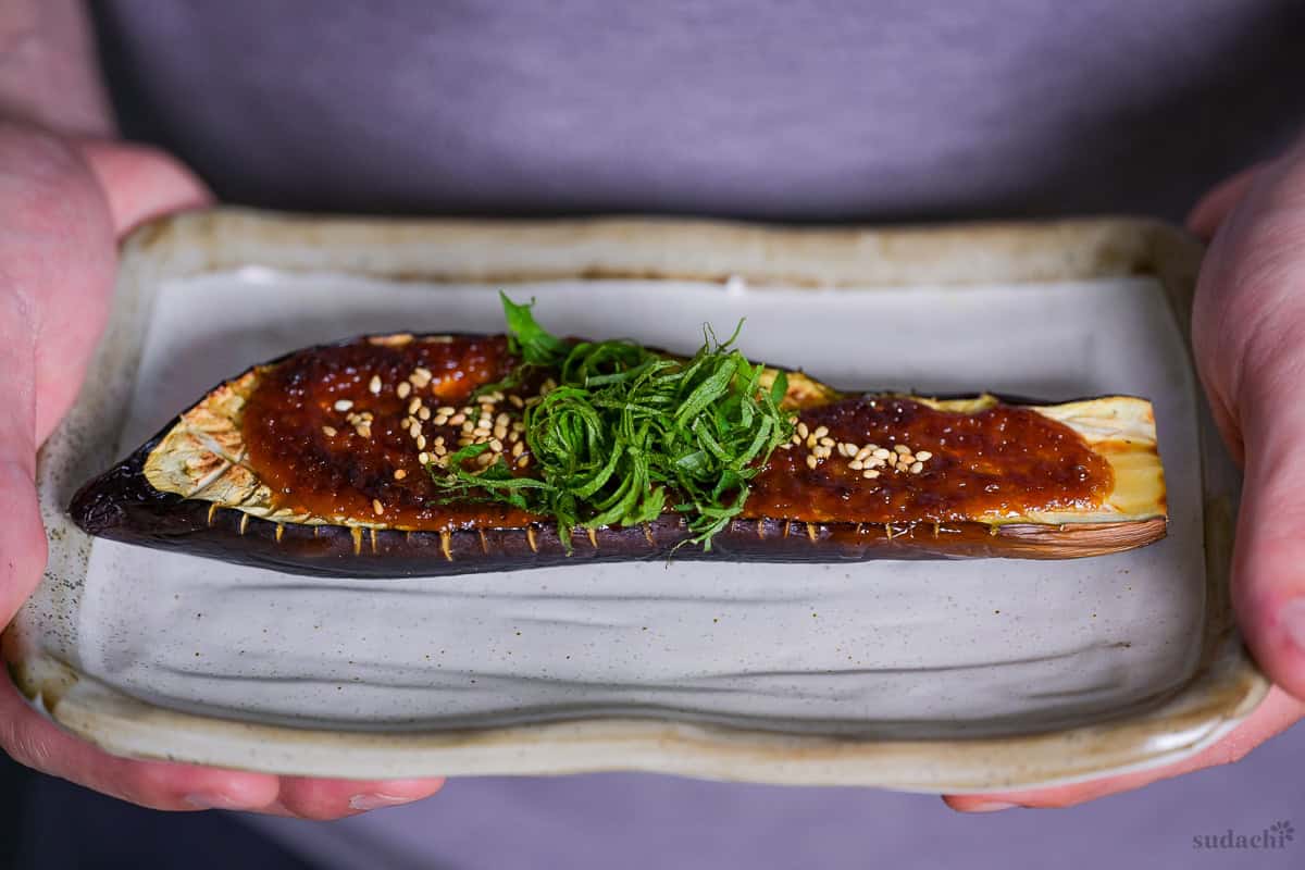 Yuto Omura holding nasu dengaku topped with toasted white sesame seeds and shredded shiso leaves on a cream-colored rectangular plate