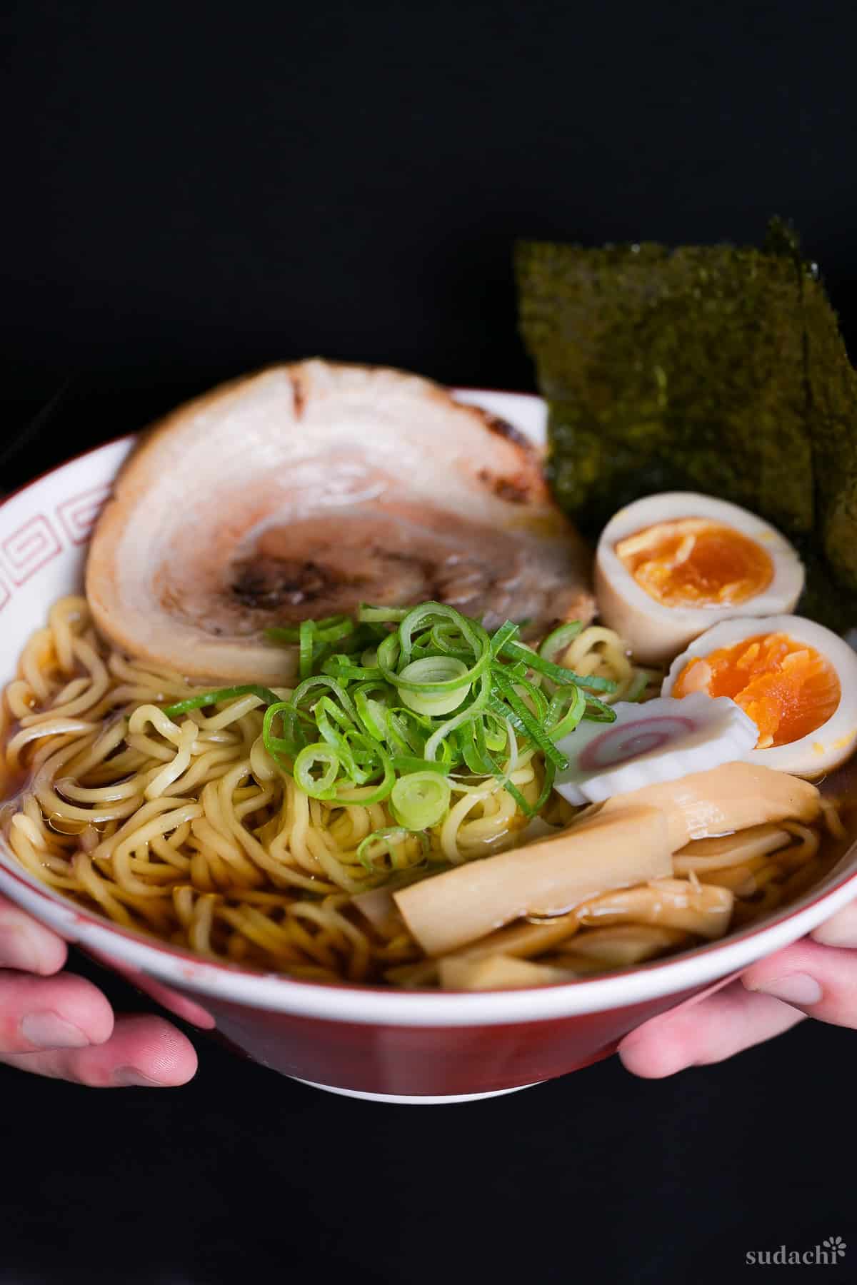 Yuto Omura holding up a bowl of homemade shoyu ramen topped with chopped green onions, menma, ramen eggs, narutoamaki fishcakes, nori, and homemade chashu served in a red and white ramen bowl