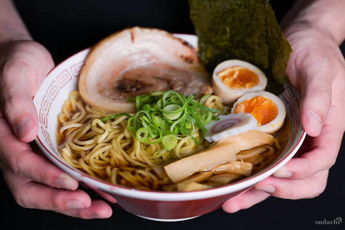 Yuto Omura holding up a bowl of homemade shoyu ramen topped with chopped green onions, menma, ramen eggs, narutoamaki fishcakes, nori, and homemade chashu served in a red and white ramen bowl