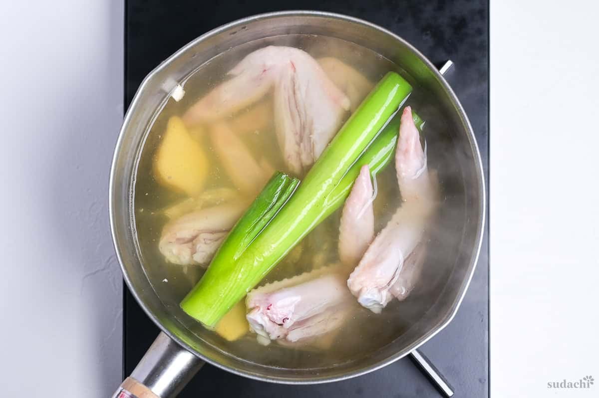 Shoyu ramen broth base with Japanese leek, chicken wings and ginger root visible, simmering in a pot on the stove top