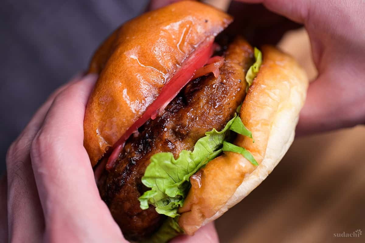 Two hands holding a teriyaki burger with lettuce, tomato and pickled ginger