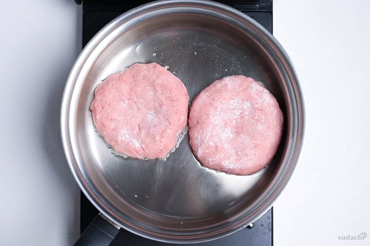 two ground pork patties frying in a stainless steel frying pan