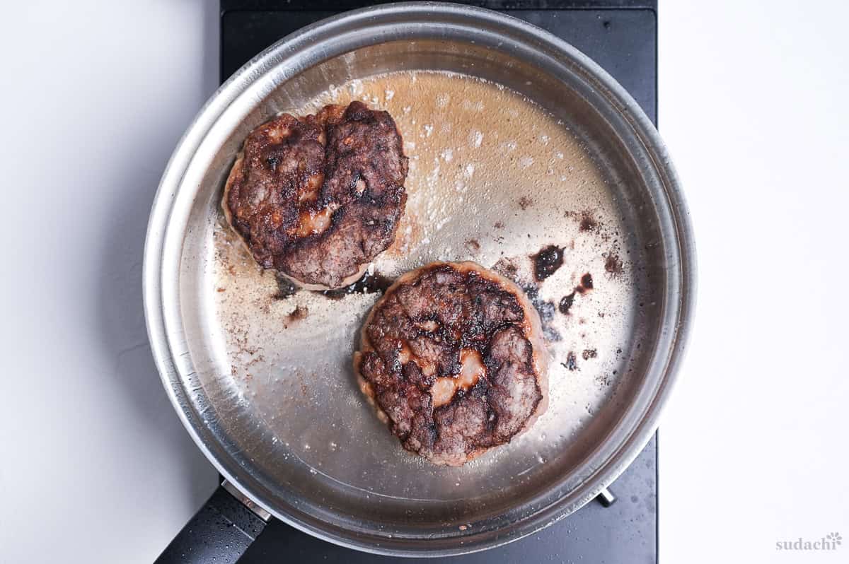 two browned ground pork patties frying in a stainless steel pan