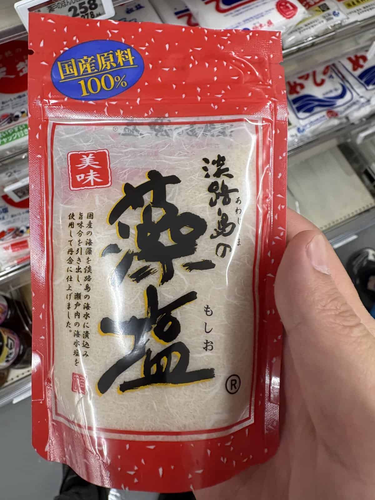 Red resealable pouch of Awajishima no Moshio, a Japanese seaweed-infused sea salt from Awaji Island, displayed on a Japanese supermarket shelf with a "100% domestic ingredients" badge.