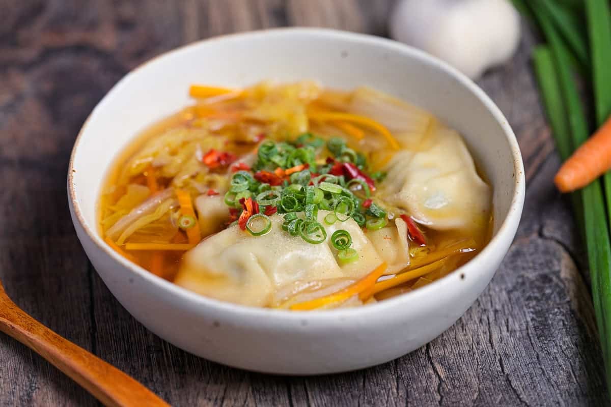 Close up of homemade gyoza soup topped with chopped green onions, chili and white pepper in a cream speckled bowl with garlic, garlic chives and a carrot in the background.