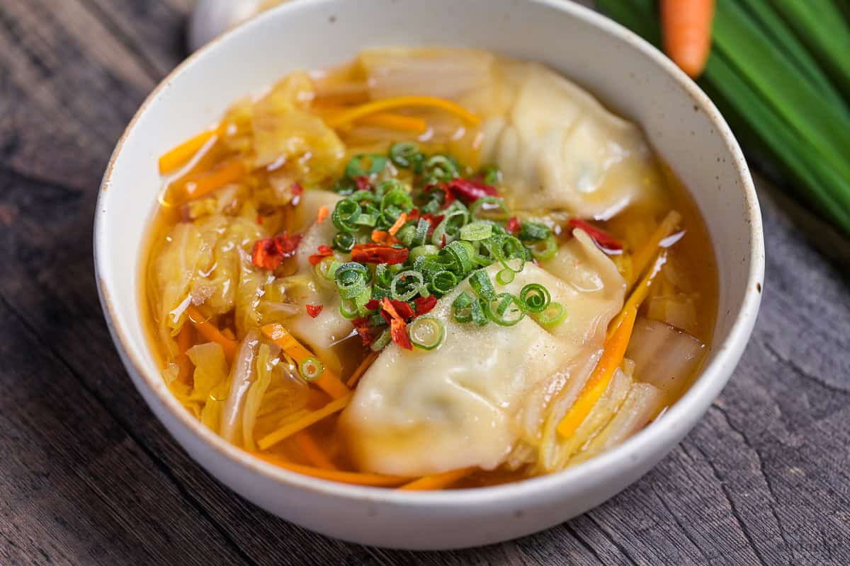 Close up of homemade gyoza soup topped with chopped green onions, chili and white pepper in a cream speckled bowl with garlic, garlic chives and a carrot in the background.
