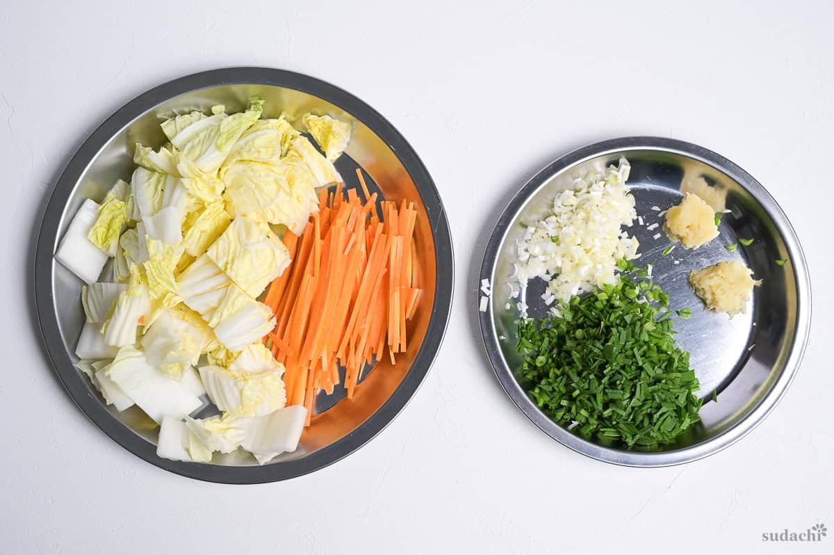 Ingredients for gyoza soup prepared and arranged on steel prep plates. Includes roughly chopped Napa cabbage, julliened carrot, finely diced Japanese leek and garlic chives, and grated ginger and garlic.