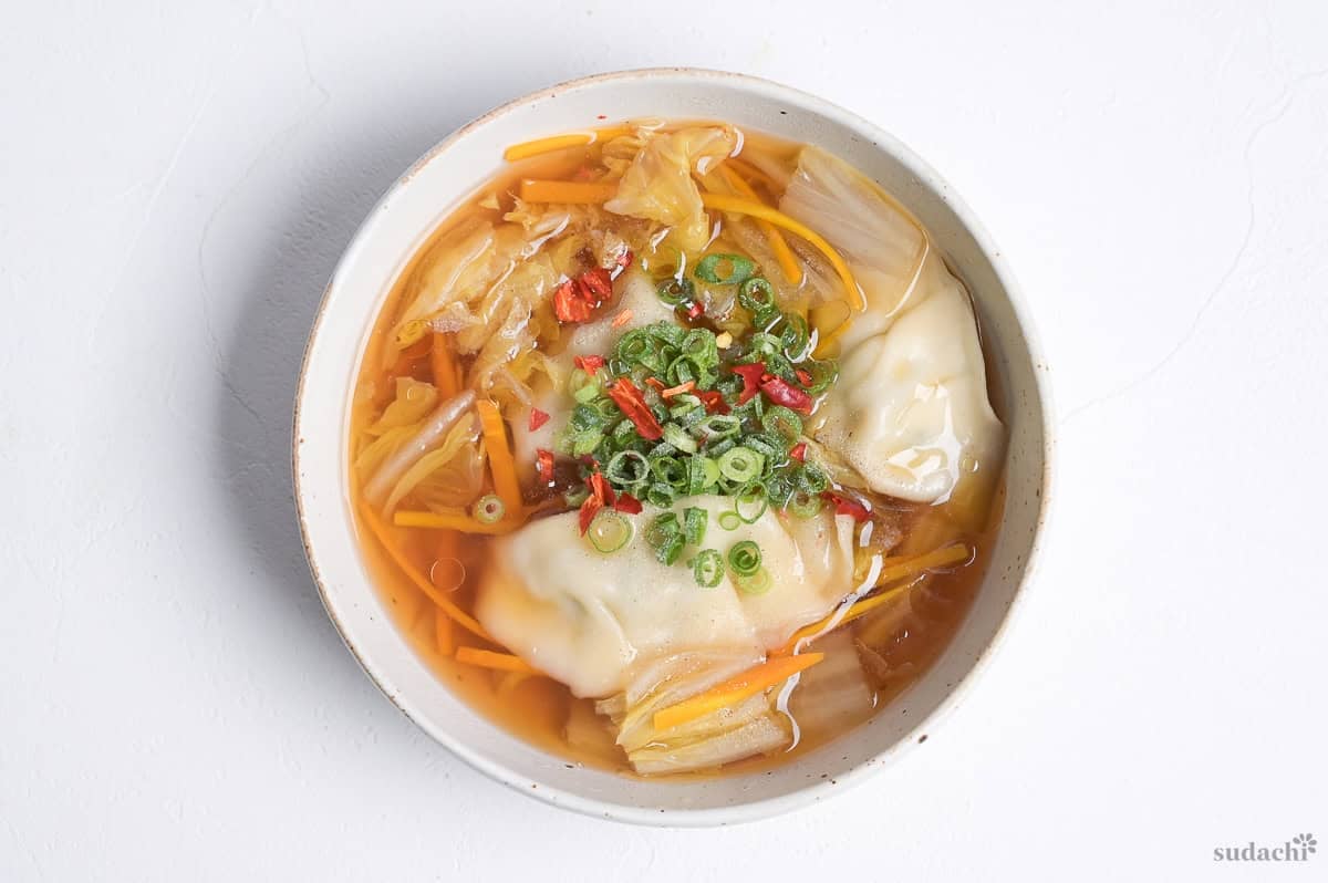 gyoza soup topped with chopped green onions, dry red chili and white pepper in a cream bowl