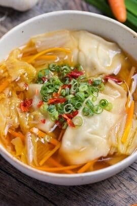 Close up of homemade gyoza soup topped with chopped green onions, chili and white pepper in a cream speckled bowl with garlic, garlic chives and a carrot in the background.