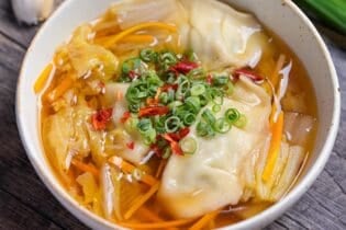 Close up of homemade gyoza soup topped with chopped green onions, chili and white pepper in a cream speckled bowl with garlic, garlic chives and a carrot in the background.