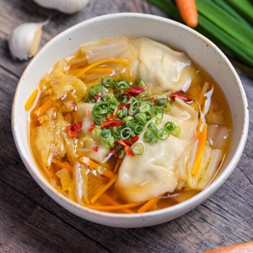 Close up of homemade gyoza soup topped with chopped green onions, chili and white pepper in a cream speckled bowl with garlic, garlic chives and a carrot in the background.