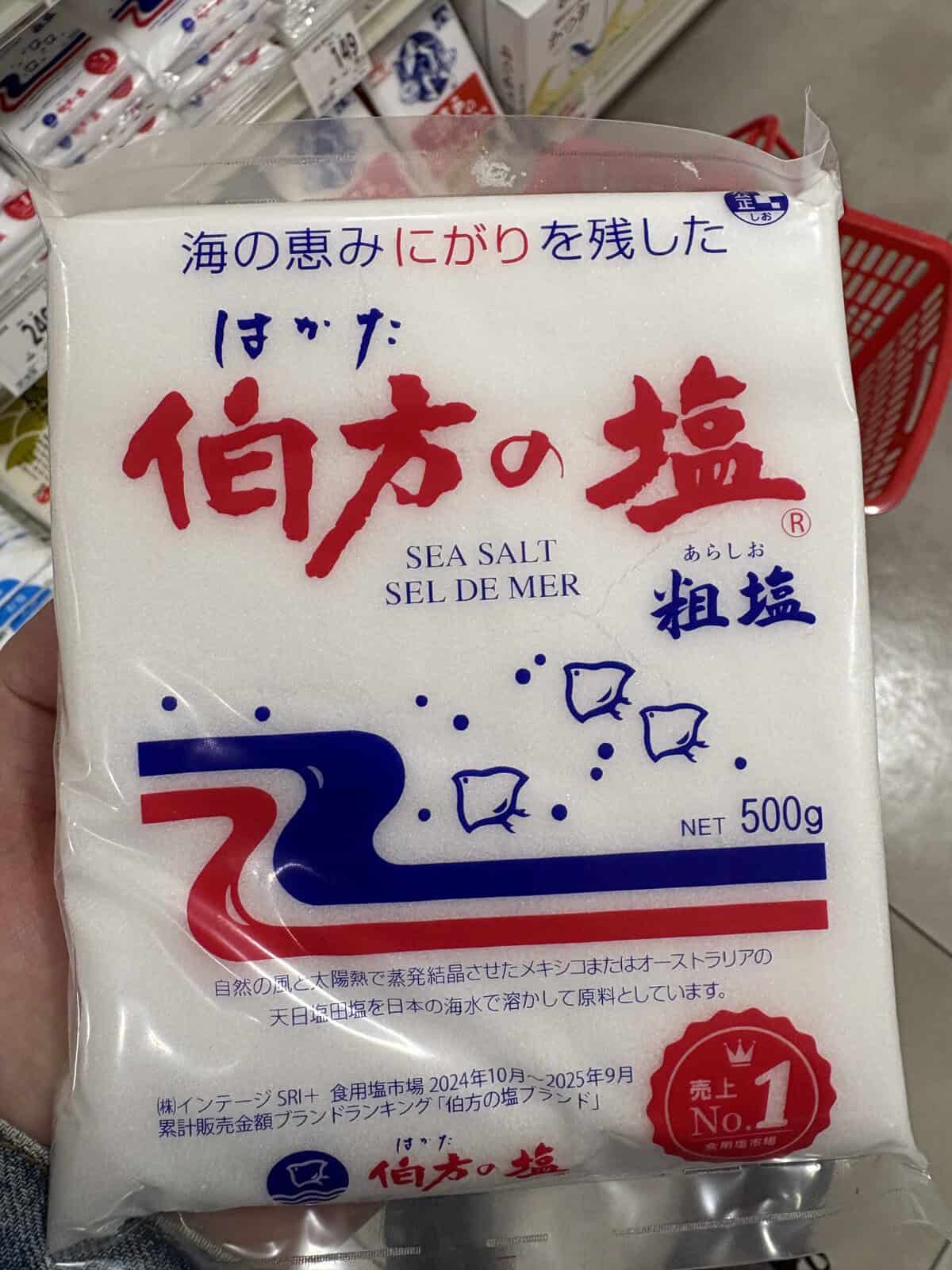 A hand holding a 500g bag of Hakata no Shio (伯方の塩) coarse sea salt in a Japanese supermarket