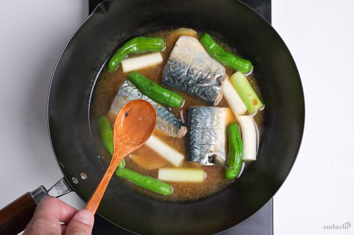 A wooden spoon with broth hovering over miso simmered mackerel in a pan (with shishito peppers and Japanese leek pieces)