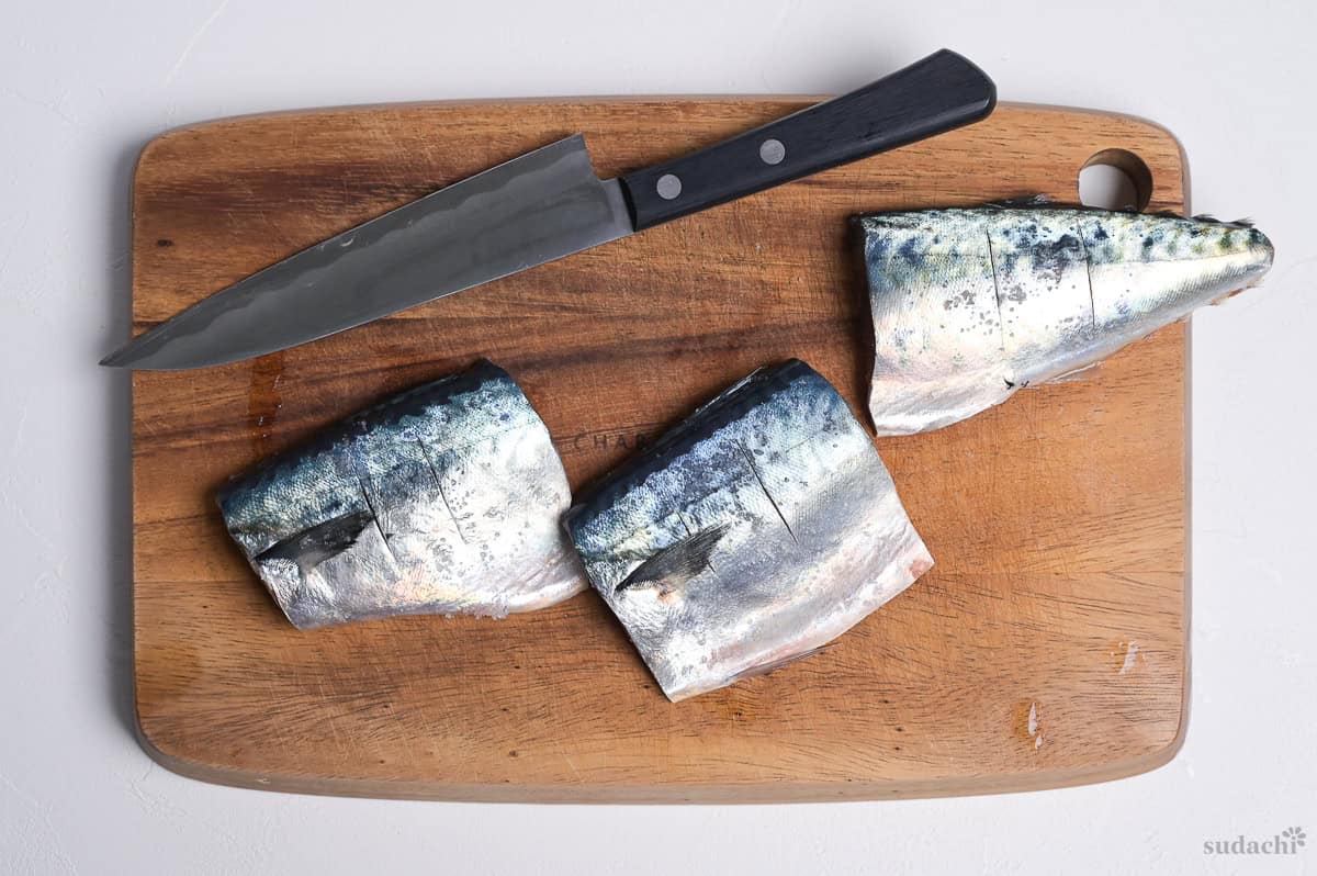 Three mackerel fillets on a wooden cutting board with vertical incisions made across them with a knife