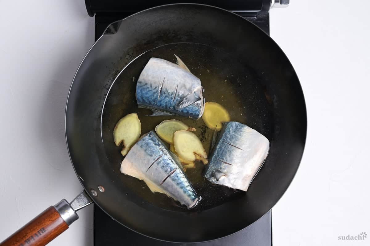 Three mackerel fillets simmering in a pan with broth and ginger root slices.