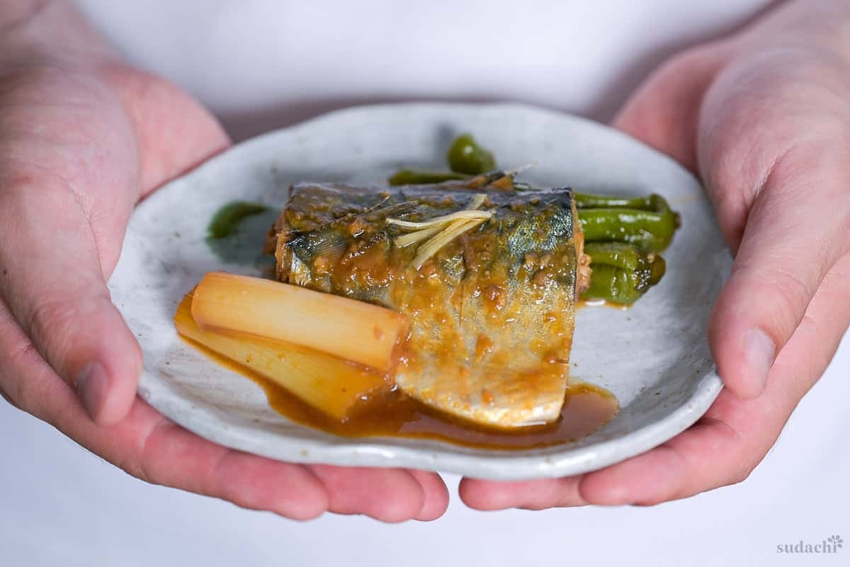 Yuto Omura holding up miso simmered mackerel on a plate with Japanese leek and shishito peppers.