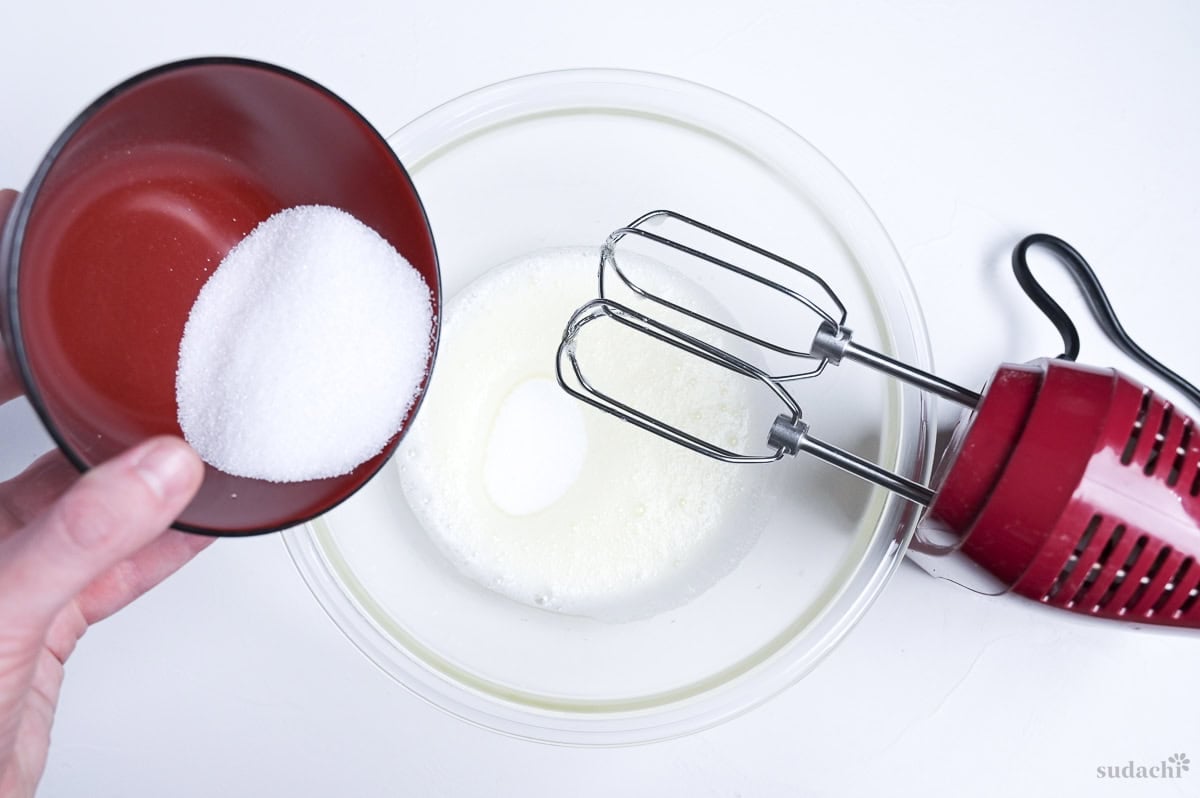 pouring half of sugar into egg whites to make a soft meringue for soufflé cheesecake