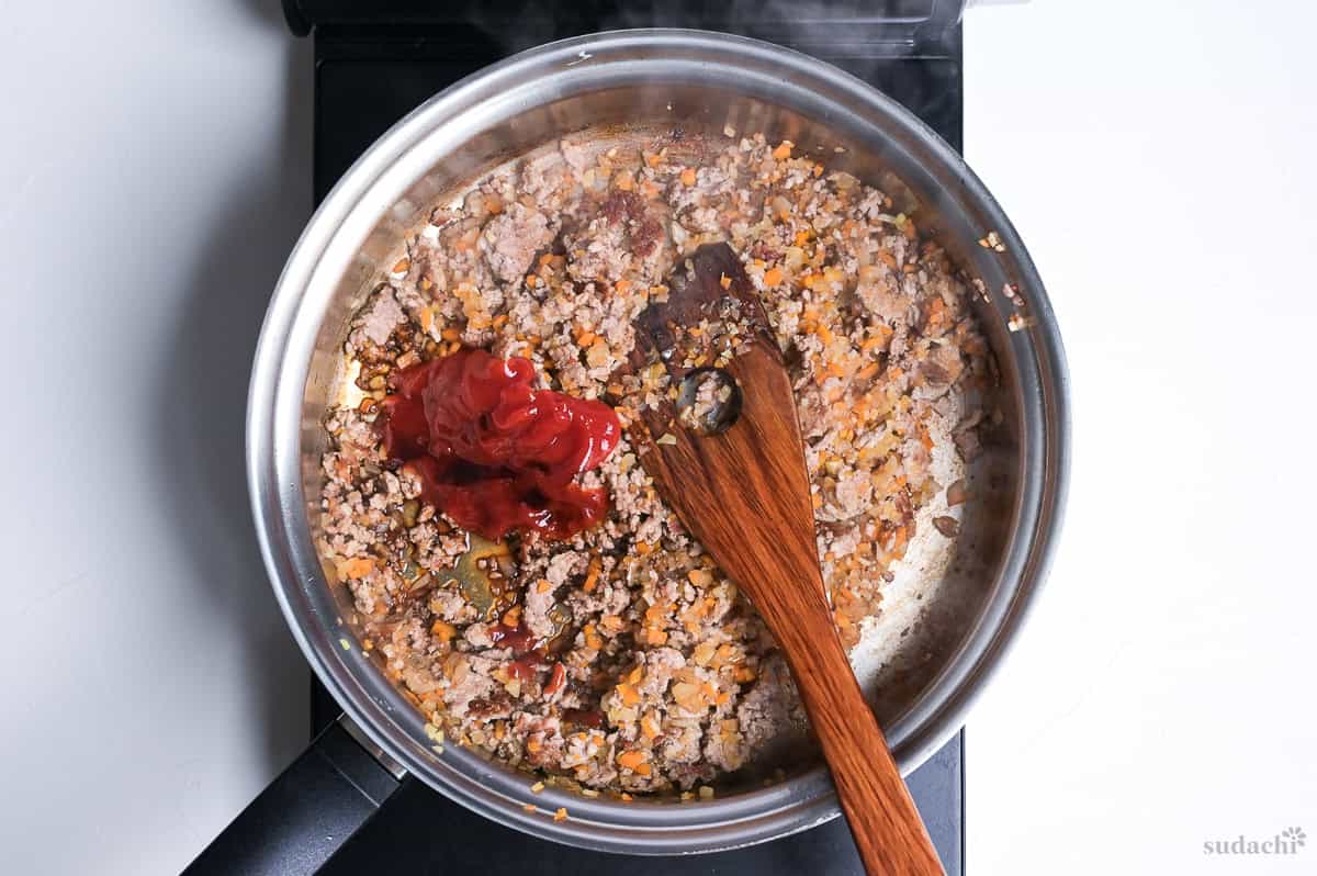 adding ketchup and Worcestershire sauce to spaghetti meat sauce in a stainless steel pan