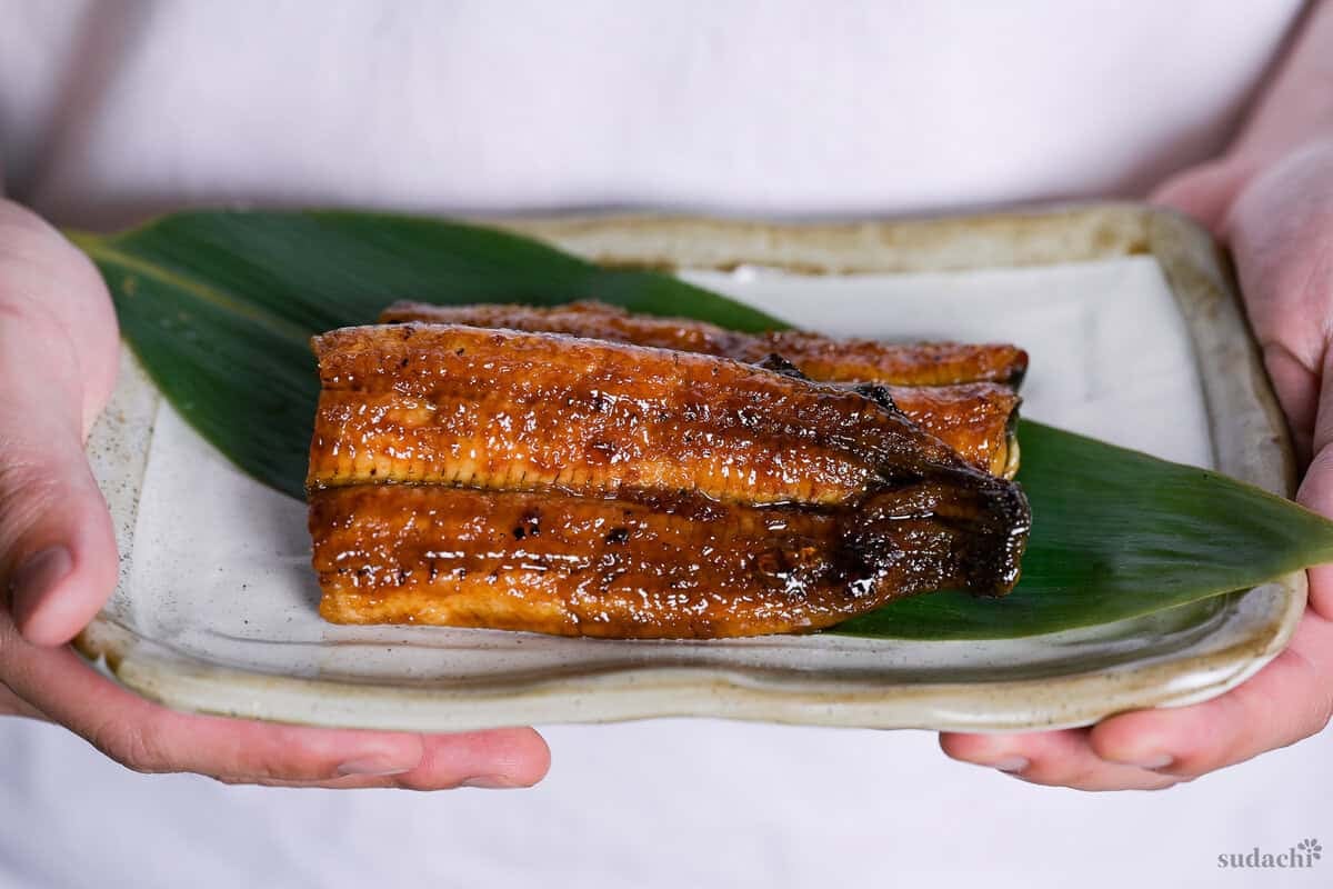 Yuto Omura holding up homemade unagi kabayaki served on a bamboo leaf on a cream rectangular plate with light brown rim