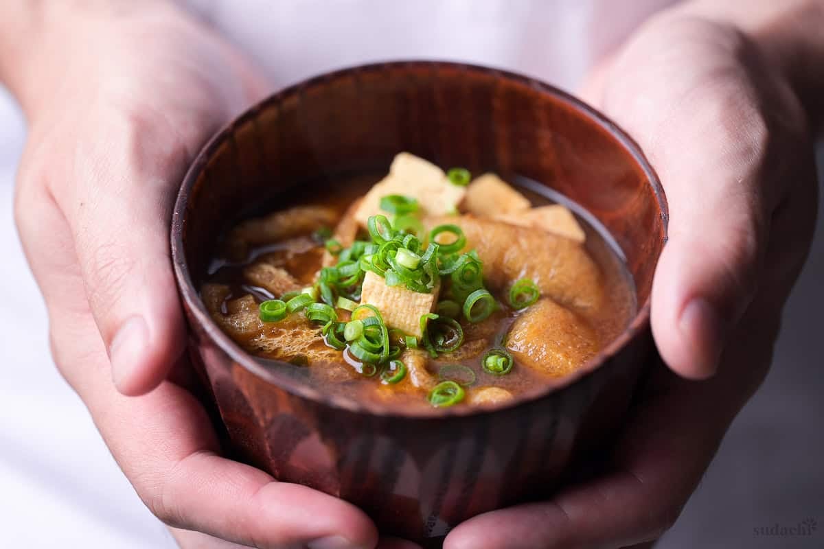Yuto Omura holding red miso soup (akadashi) topped with chopped green onions in a dark wooden Japanese soup bowl