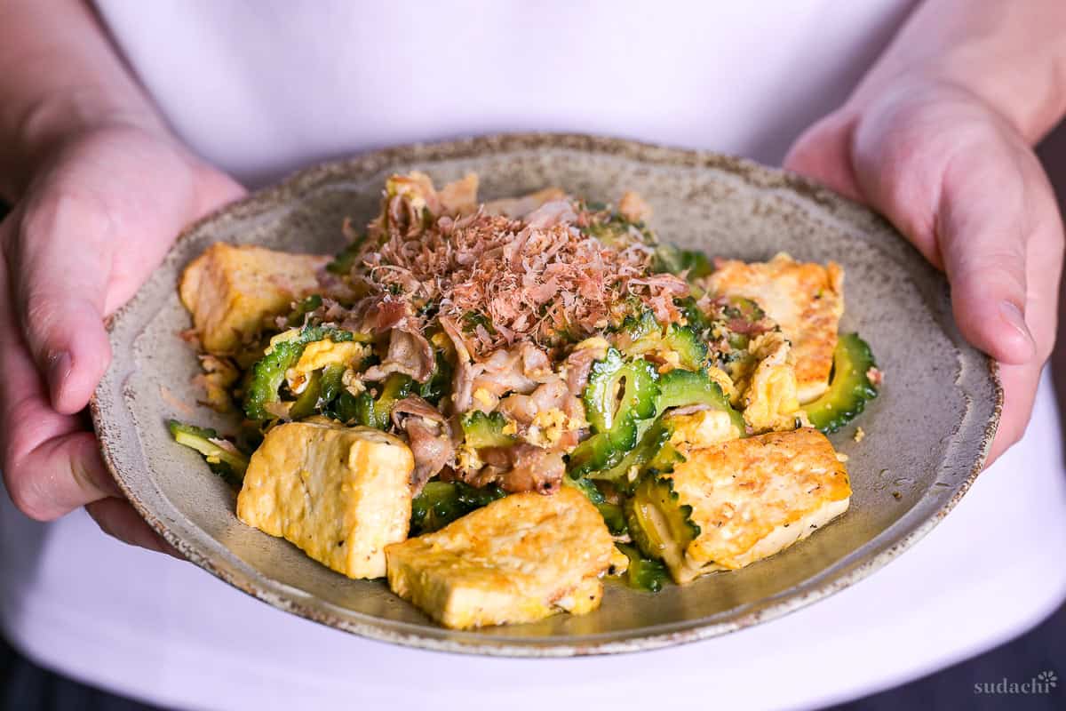 Yuto Omura holding up Goya Champuru sprinkled with bonito flakes on a brown plate