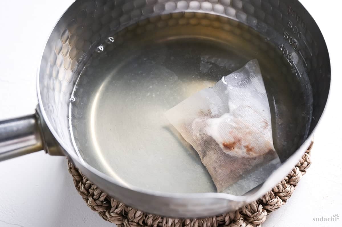 Dashi packet simmered in hard water in a saucepan on a Japanese style woven trivet.