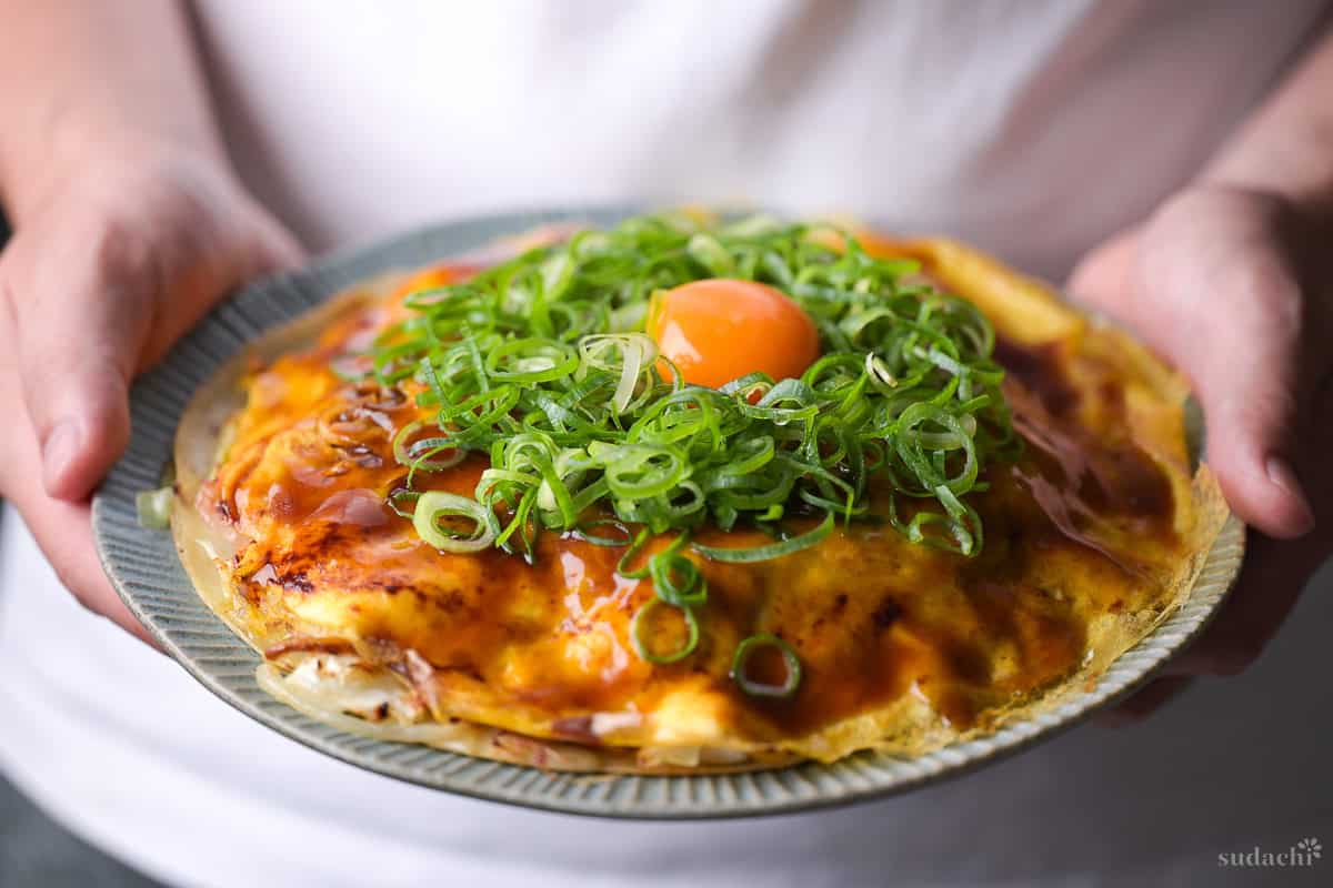 Yuto Omura holding up Hiroshima Style Okonomiyaki topped with green onions and an egg yolk on a plate