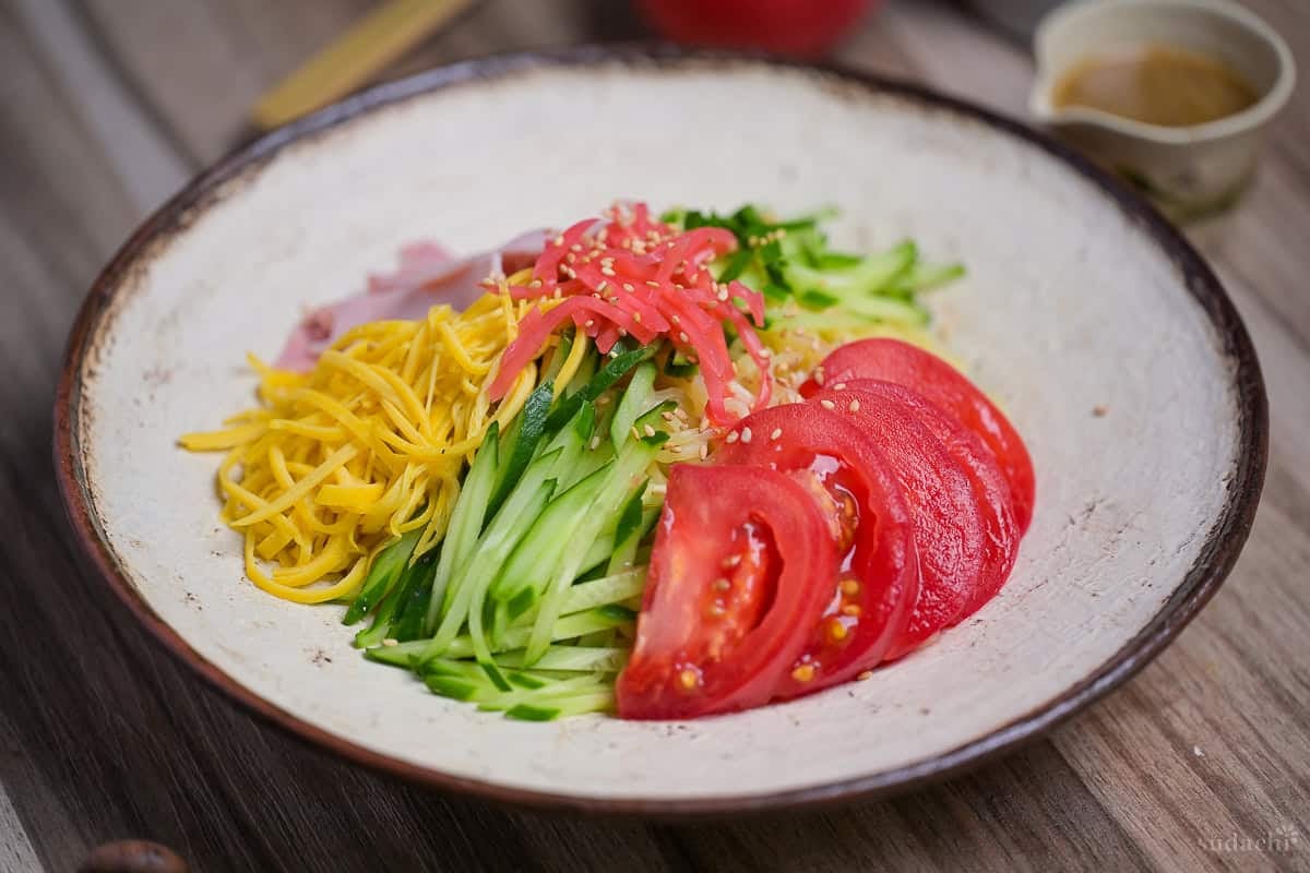 Hiyashi chuka topped with shredded egg crepe, cucumber, tomato, ham, benishoga pickles and toasted white sesame seeds served on a cream textured plate with brown rim.