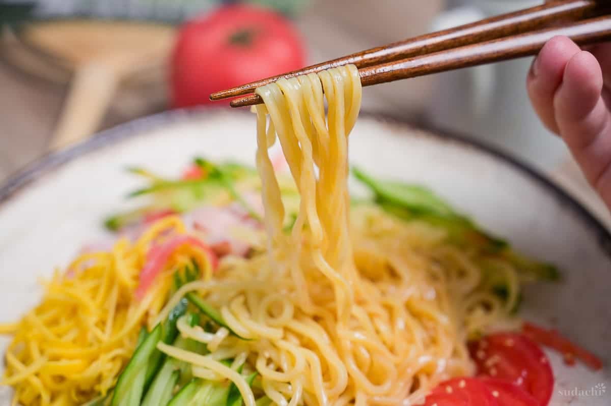 Close up of wooden chopsticks holding up noodles from hiyashi chuka.
