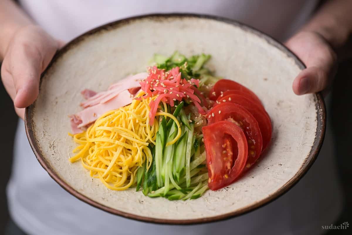 Yuto Omura holding up hiyashi chuka topped with shredded egg crepe, cucumber, tomato, ham, benishoga pickles and toasted white sesame seeds served on a cream textured plate with brown rim.