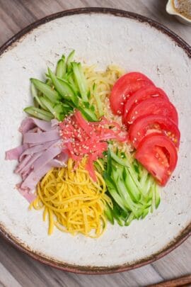 Hiyashi chuka topped with shredded egg crepe, cucumber, tomato, ham, benishoga pickles and toasted white sesame seeds served on a cream textured plate with brown rim top down view.