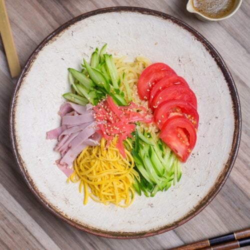 Hiyashi chuka topped with shredded egg crepe, cucumber, tomato, ham, benishoga pickles and toasted white sesame seeds served on a cream textured plate with brown rim top down view.