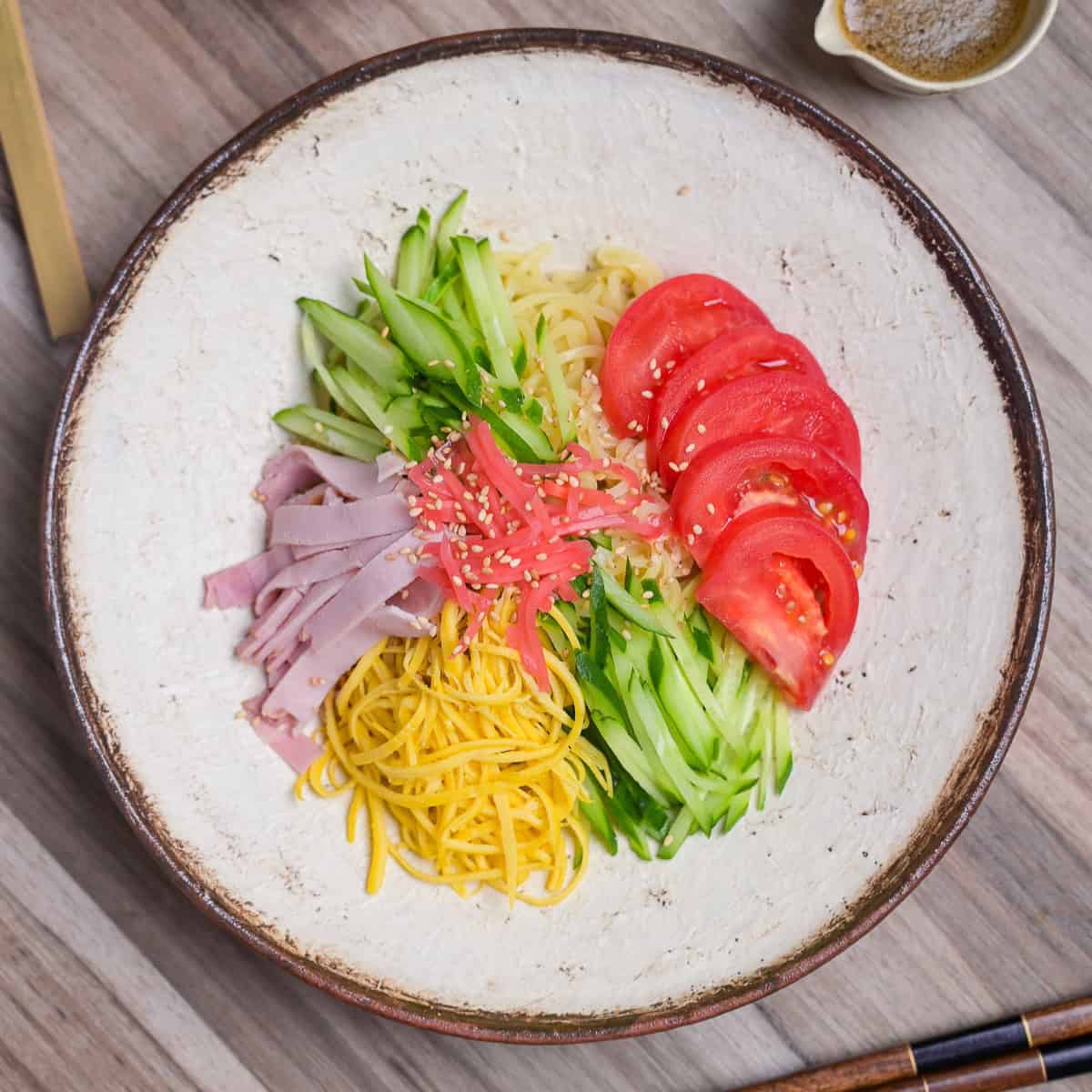Hiyashi chuka topped with shredded egg crepe, cucumber, tomato, ham, benishoga pickles and toasted white sesame seeds served on a cream textured plate with brown rim top down view.