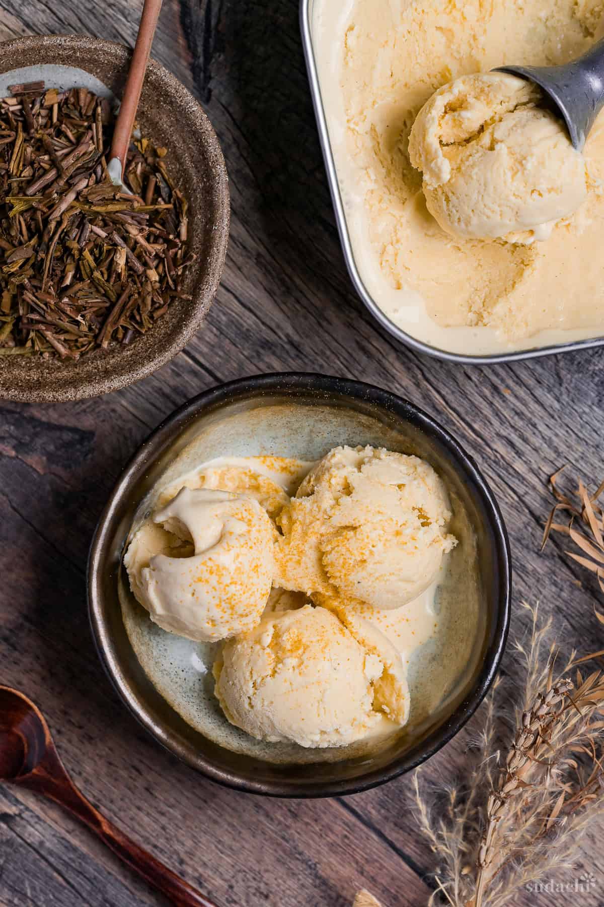 Three scoops of homemade hojicha ice cream in a bowl next to loose hojicha tea in a brown dish, and hojicha ice cream in a steel tub with scoop scooping