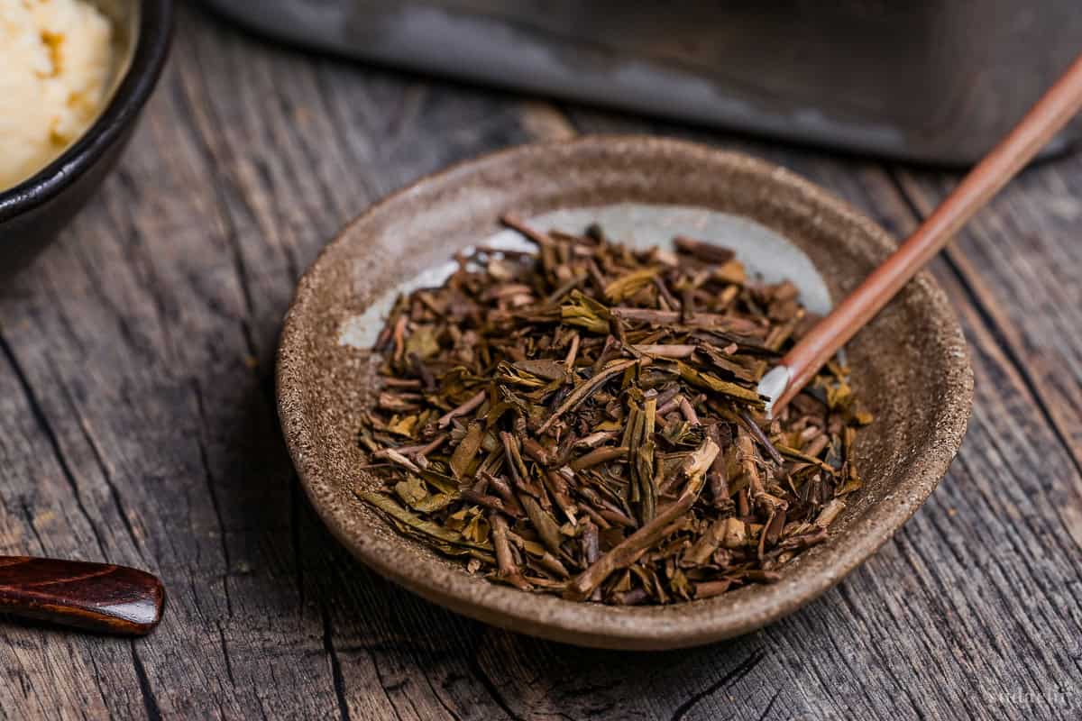 Loose hojicha tea in a brown dish with small ceramic spoon
