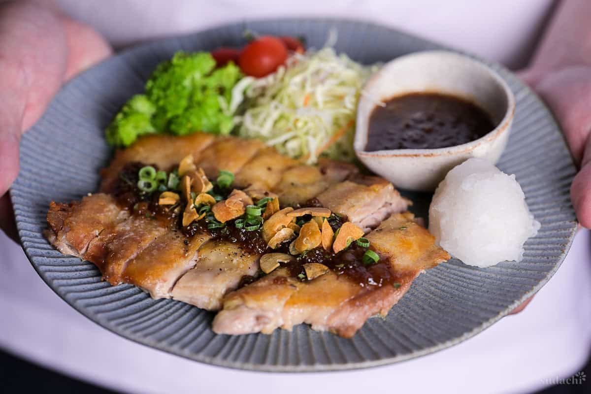 Yuto Omura holding up Japanese style pan fried chicken steak with homemade sauce and crispy garlic chips on a blue plate