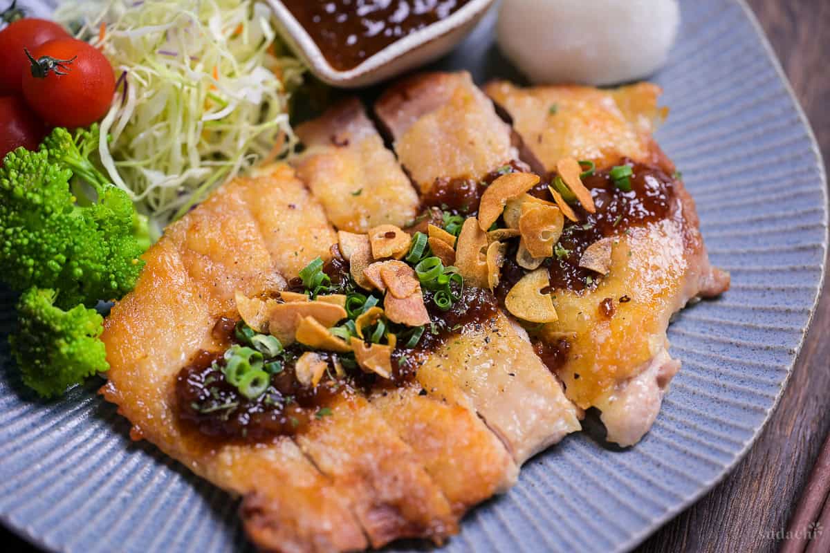 Japanese style pan fried chicken steak with homemade sauce and crispy garlic chips on a blue plate close up
