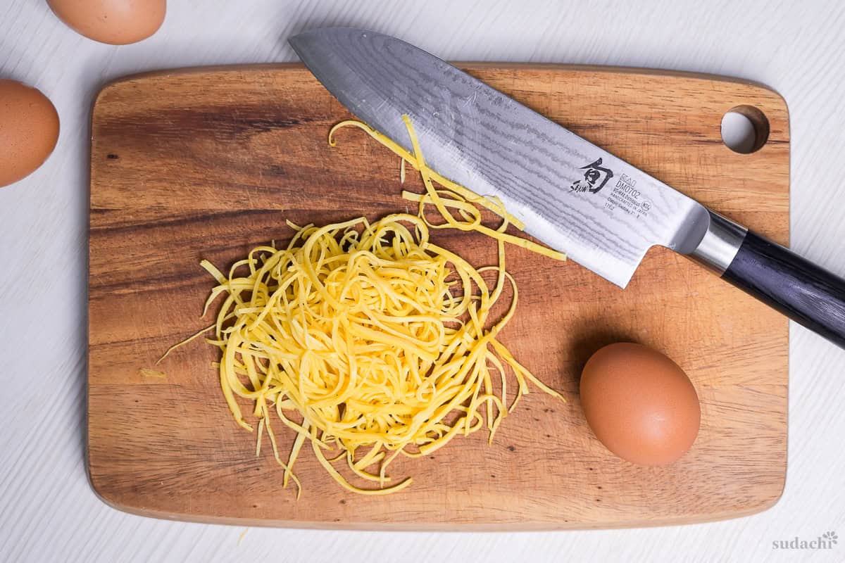 Homemade Kinshi Tamago (Japanese shredded egg crepe) on a wooden cutting board next to a Japanese knife and brown egg