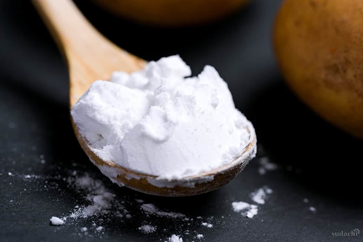 A scoop of potato starch (katakuriko) on a light wooden spoon on a black cutting board with potatoes in the background