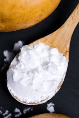 A scoop of potato starch (katakuriko) on a light wooden spoon on a black cutting board with potatoes in the background