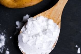 A scoop of potato starch (katakuriko) on a light wooden spoon on a black cutting board with potatoes in the background