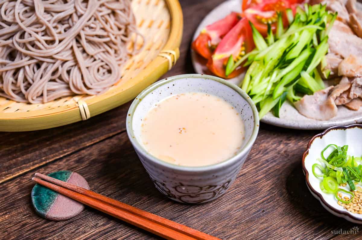 Sesame dipping sauce for soba noodles in a small gray dipping bowl, with tomato wedges, julienned cucumber and seared pork on a plate in the background