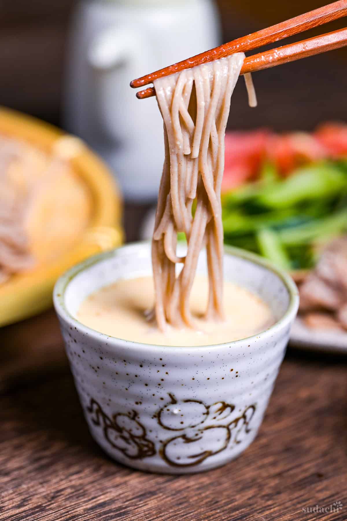 Wooden chopsticks holding soba noodles dipped in homemade sesame dipping sauce in a gray mottled ceramic dipping bowl