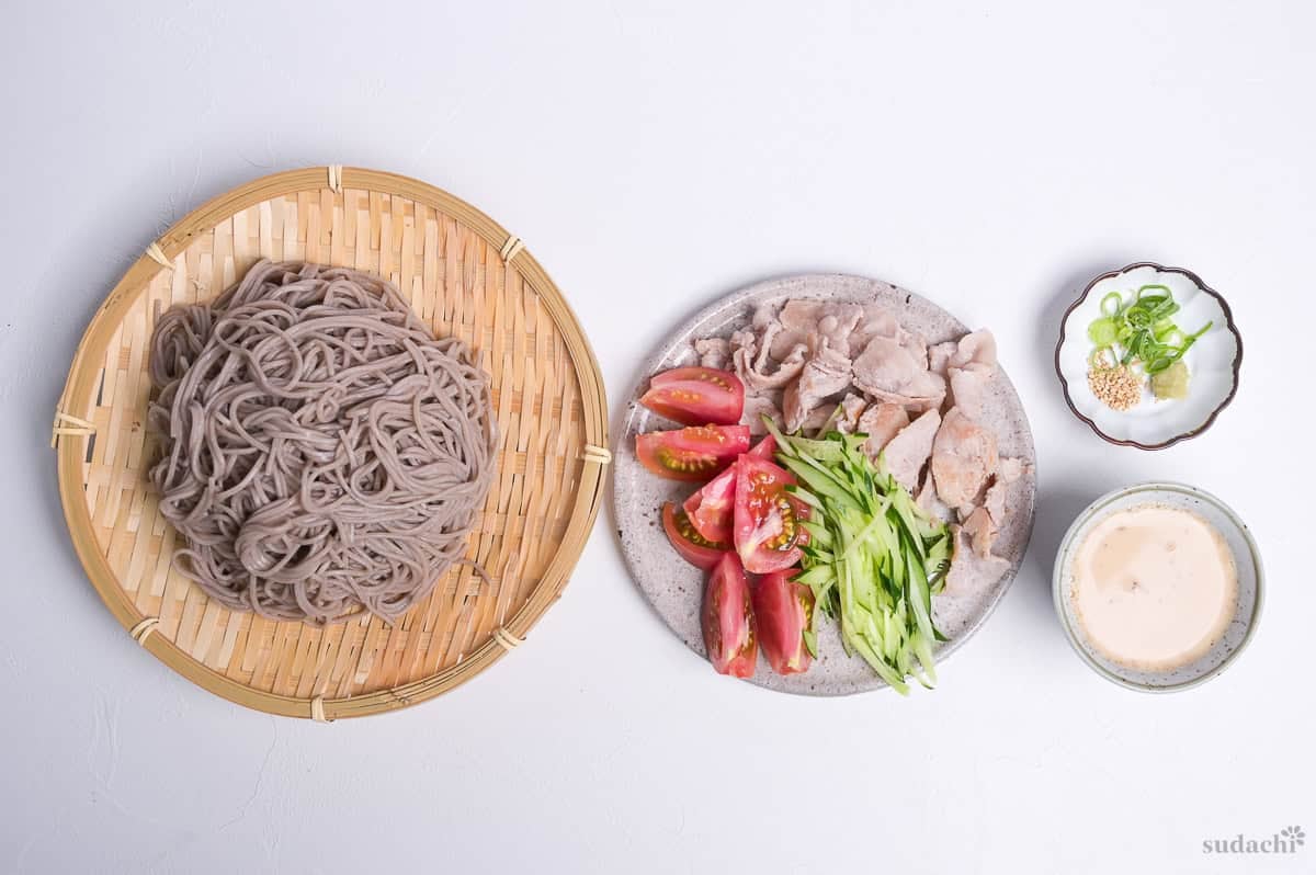 cooked and chilled soba noodles on a bamboo tray (zaru), next to seared pork, tomato wedges and julienned cucumber on a plate, next to sesame sauce and a small plate with sesame seeds, chopped green onions and wasabi for toppings.