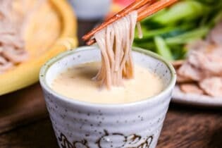 Sesame dipping sauce for soba noodles in a small gray dipping bowl, with tomato wedges, julienned cucumber and seared pork on a plate in the background