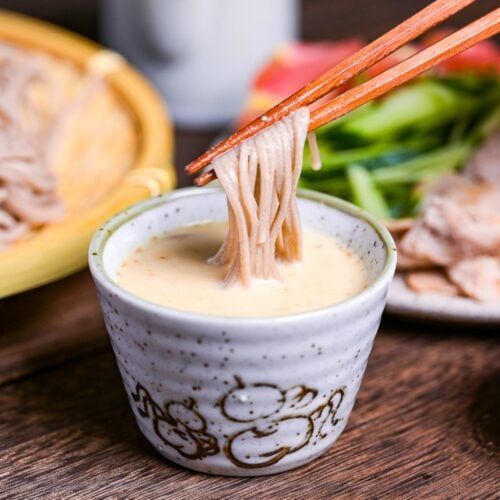 Sesame dipping sauce for soba noodles in a small gray dipping bowl, with tomato wedges, julienned cucumber and seared pork on a plate in the background