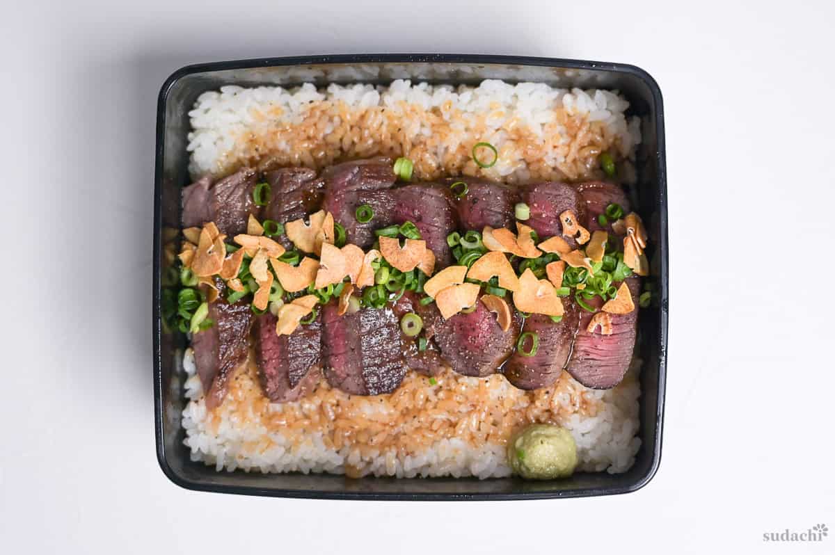 Steak Jyu with chopped green onions, crispy garlic chips and a blob of wasabi in a black jubako (box)